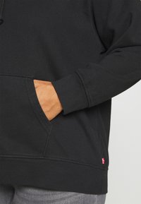 La mano di una persona poggiata all'interno della tasca anteriore di un felpa nera con un piccolo etichetta rossa del marchio sul lato inferiore destro.