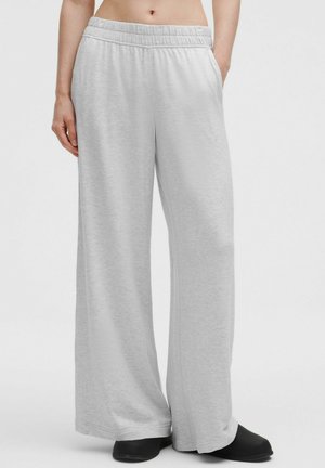 Lichtgrijze broek met wijde pijpen en elastische tailleband, gedragen door een persoon die staat met handen in de zakken en zwarte instapschoenen.