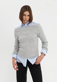 Maglione grigio a costine con design off-the-shoulder sopra una camicia colletto azzurra, abbinato a pantaloni neri. Tessuto morbido e silhouette aderente.