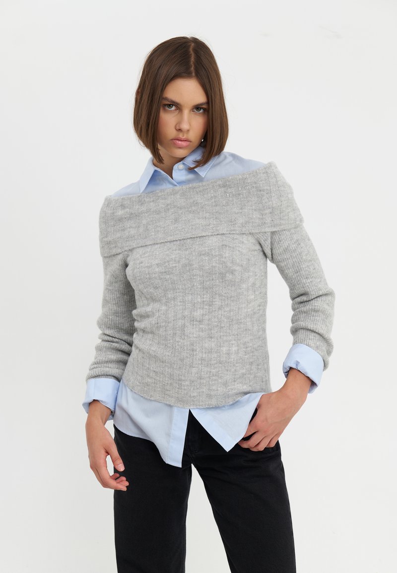 Maglione grigio a costine con design off-the-shoulder sopra una camicia colletto azzurra, abbinato a pantaloni neri. Tessuto morbido e silhouette aderente.