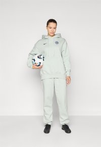 Lys grå hettegenser med lomme foran og logo, kombinert med matchende joggebukser. Svart joggesko og en mønstret fotball holdes.