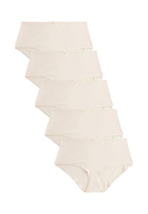 Femme aux longs cheveux bruns portant un soutien-gorge beige et une culotte taille haute assortie, debout devant un fond clair uni, souriant légèrement.