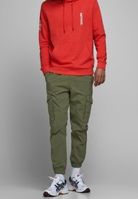 Röd hoodie med logotyp, dragsnören och framficka. Olivgröna cargo-byxor med elastiska fållar. Vita sneakers med svarta detaljer.