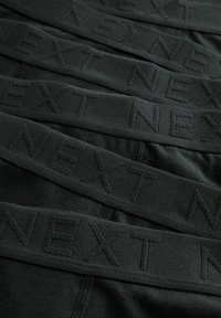 Banda elastica neagră cu inscripția „NEXT” embosată, stratificată peste un fundal de țesătură netedă. Textură mată fără modele vizibile.