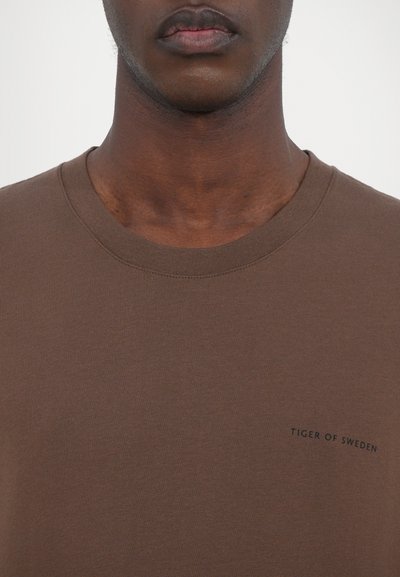Brown cotton t-shirt with a crew neckline, featuring a small logo text on the chest reading "TIGER OF SWEDEN." Smooth texture, standard fit.