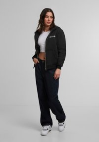 Zwarte hoodie met ritssluiting en een voorzak, gecombineerd met donkere wijde jeans en witte sneakers met accenten. Soepel textuur.