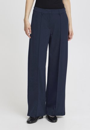 Osoba nosící tmavě modré široké kalhoty s předními švy a černé boty, stojící před jednobarevným světlým pozadím.