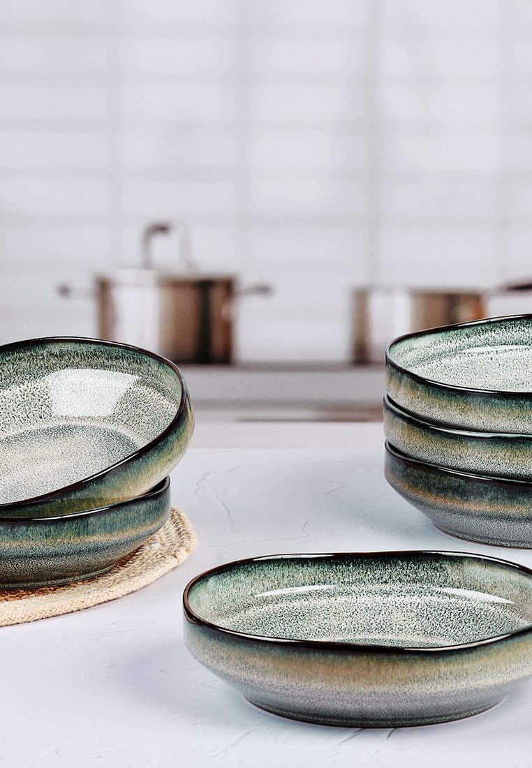 Keramische Schalen mit abgerundeten Kanten, die mit einer gesprenkelten grün-grauen Oberfläche versehen sind und auf einem runden gewebten Mattenstapel liegen.