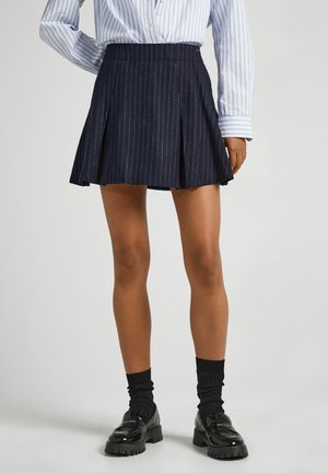 Vrouw die een marineblauwe geplooide minirok met krijtstreep, lichtblauw gestreept overhemd, zwarte sokken en zwarte chunky loafers draagt tegen een effen achtergrond.