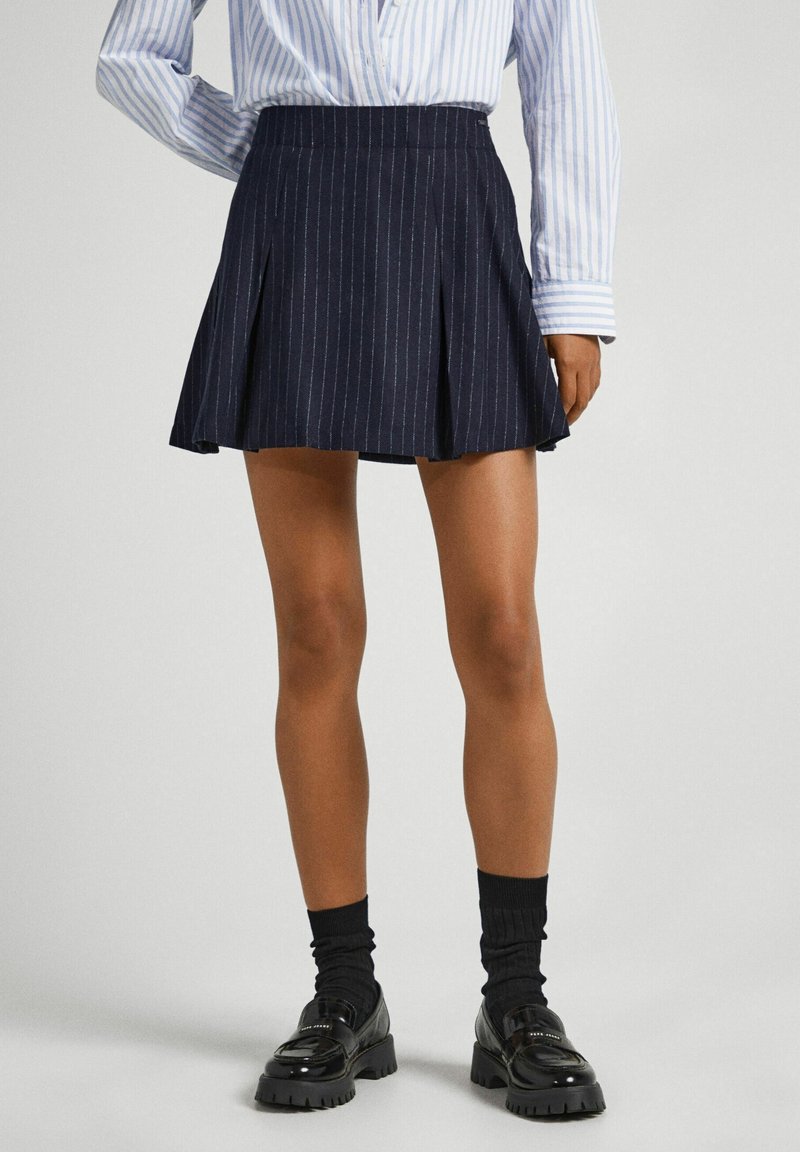 Femme portant une mini-jupe plissée bleu marine à fines rayures, une chemise rayée bleu clair, des chaussettes noires et des mocassins noirs épais sur un fond uni.