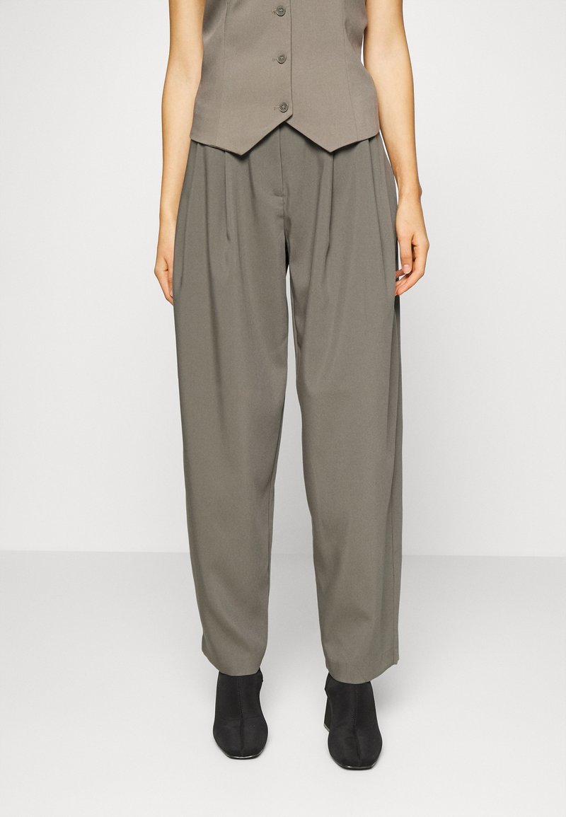 Femme portant un pantalon large plissé vert olive, un gilet assorti et des bottines noires, debout devant un fond blanc uni.