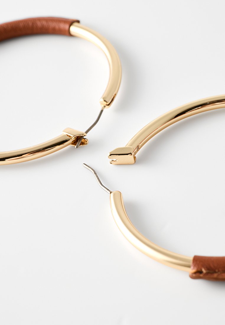 Boucles d'oreilles en ton or avec un design lisse et courbé et un accent en cuir marron. Présentent une fermeture à cliquet et une silhouette élégante.