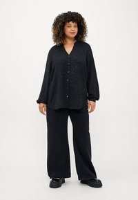 Zwarte knoopshirt met V-hals, lange mouwen en een relaxte pasvorm, met een subtiel glinsteringen patroon, gecombineerd met wijde zwarte broeken.