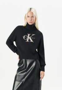 Zwarte gebreide coltrui met een groot crèmekleurig "CK"-logo en de tekst "Calvin Klein", gecombineerd met een zwarte leren kokerrok.