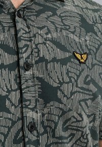 Groene button-up shirt met een gestructureerd abstract patroon in het wit, met een klein geel geborduurd logo op de borst en zwarte knopen.