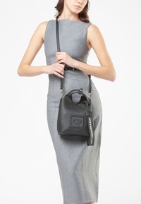 Donna in un abito grigio aderente senza maniche, che tiene una borsa a secchiello in pelle nera con una tracolla e un'etichetta che dice "SONO UNA BORSA RESPONSABILE."