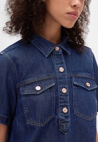 Jeansskjorta med korta ärmar, mörkblå färg, två bröstfickor och kopparfärgade tryckknappar. Har klassisk krage och texturerat tyg.