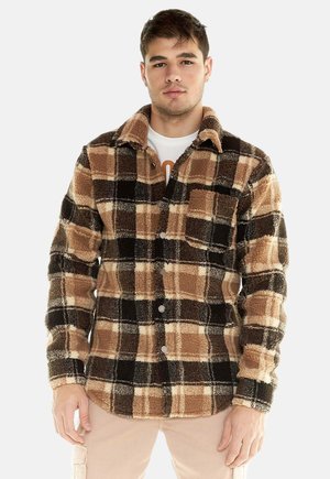 Un homme porte une veste polaire à carreaux bruns, noirs et crème par-dessus une chemise blanche, sur un fond blanc uni.