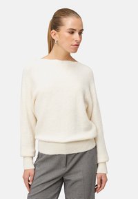 Kremowy sweter o miękkiej teksturze z szerokim dekoltem i d�ługimi, mankietowymi rękawami, w zestawie z szarymi spodniami w kratę.