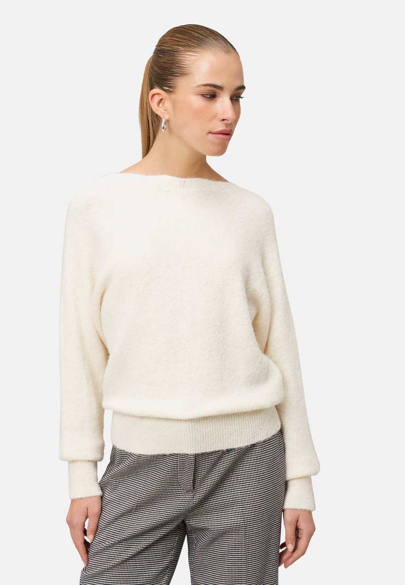 Kremowy sweter o miękkiej teksturze z szerokim dekoltem i długimi, mankietowymi rękawami, w zestawie z szarymi spodniami w kratę.