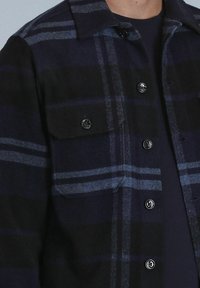 Chemise en flanelle de couleur bleu marine foncé et noir, avec des rayures en tartan bleu. Elle possède un devant boutonné et deux poches poitrine avec fermetures à pression.