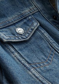 Gros plan sur un tissu en denim avec une teinte bleue, des coutures contrastées, et un bouton argenté embossé du texte « Authentic Next Dude 1982 ».