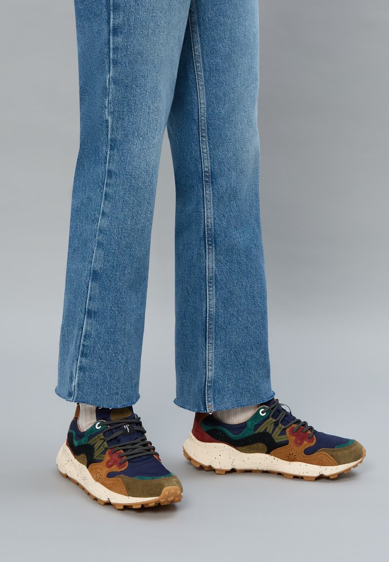 Flerfärgade sneakers i marinblått, grönt, rött och brunt, med en fläckig sula och texturerade tygdetaljer, kombinerade med blå jeans.