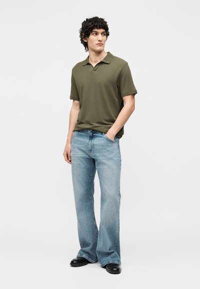 Jeune homme aux cheveux bouclés portant un polo vert olive, un jean bleu clair à jambes larges et des chaussures noires, debout avec une main dans la poche.