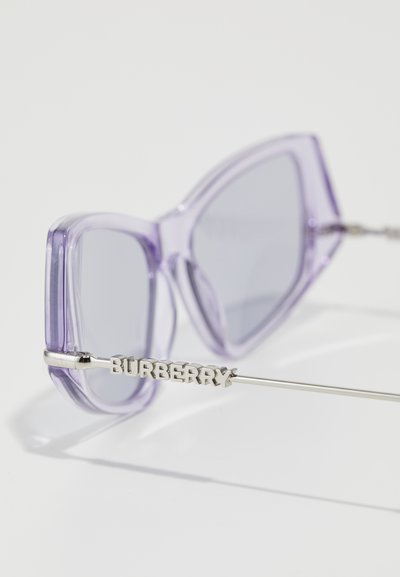 Violetti läpinäkyvät aurinkolasit, joissa on kulmikas katti-silmä muotoilu, harmaat linssit ja hopeiset metalliset kehykset, joihin on merkitty brändin nimi "Burberry."