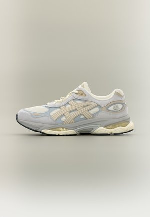 Baskets de sport blanches et beiges avec une tige en mesh et cuir, des bandes beiges, une semelle coussinée et un amorti à air visible sur fond gris.