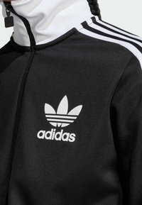 Svart jacka med vit hög krage, med tre vita ränder på axlarna och Adidas-logotyp med tre bladmönster på bröstet.