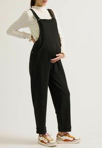 Schwangere Frau in einem schwarzen Overall mit langem Ärmel, lächelnd und sanft ihren Bauch haltend, trägt bunte Turnschuhe.