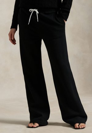 Fekete bőszárú nadrágot fehér zsinórral, fekete pulóvert és fekete szandált viselő személy bézs színű padlón áll.