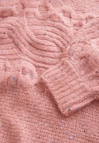 Texturerad rosa ulltröja med upphöjda kabelstickade mönster och ribbade ärmslut, med subtila färgprickar i blått och svart.
