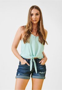 Top azzurro chiaro senza maniche con un dettaglio di nodo frontale, abbinato a pantaloni corti in denim scuro con risvolti e particolari usurati.