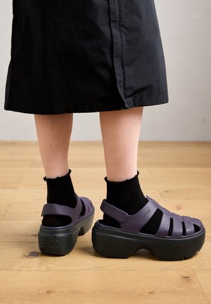 Osoba nosząca czarne skarpetki do kostek i ciemnofioletowe sandały na platformie Crocs na jasnej drewnianej podłodze, widoczna czarna spódnica.