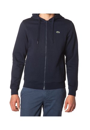 Homme portant un sweat à capuche bleu marine zippé avec poches avant et un petit logo crocodile vert sur la poitrine gauche.