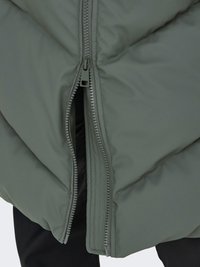 Gros plan d'une veste matelassée verte partiellement ouverte avec un pantalon noir visible en dessous.