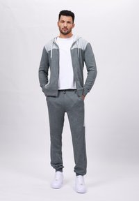 Grå zip-opp hettegenser med lysegrå detaljer, sammen med matchende joggebukser. Begge plaggene er laget av myk, teksturert stoff. Hvite sneakers.