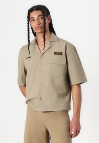 Camicia khaki a maniche corte, con bottoni e tasca, testo ricamato e etichetta patch. Realizzata in tessuto leggero, dalla superficie morbida.