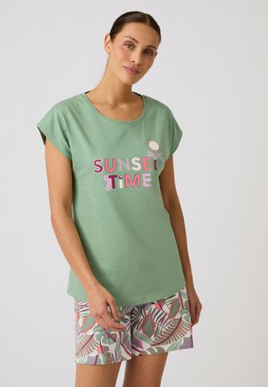 Frau trägt ein grünes T-Shirt mit dem Text "SUNSET TIME" und tropisch gemusterte Shorts, steht vor einem einfarbigen hellen Hintergrund.