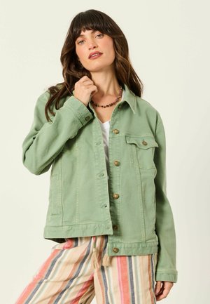 Woman with dark hair wears light green buttoned jacket and multicolored striped pants, touching jacket collar against plain background.