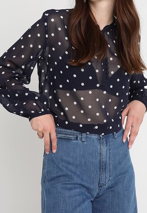 Vrouw draagt een marineblauwe, doorschijnende blouse met lange mouwen en witte polka dots en een blauw, hooggesloten spijkerbroek, handen rusten op de heupen.
