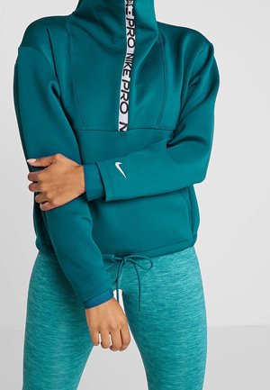 Personne portant un sweat à capuche Nike Pro bleu sarcelle avec une demi-fermeture éclair et des leggings ajustés assortis, main croisée pour tenir le bras opposé.