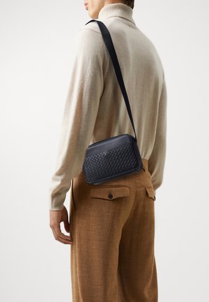 Man draagt een beige trui en bruine broek met een zwarte, gevlochten leren crossbodytas aan zijn schouder.