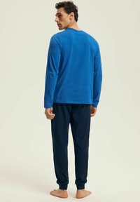 Man facing backward wearing a blue long-sleeve shirt and dark navy jogger pants, standing barefoot on a light background.