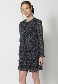 Rochie neagră din chiffon cu model floral albastru, cu mâneci lungi și tiv ondulat. Prezintă un gât rotund și pliuri la umeri.