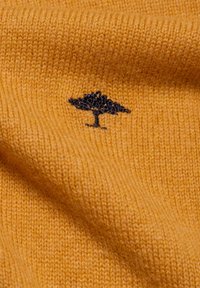 Gelbes gestricktes Material mit glatter Oberfläche, versehen mit einem dunklen gestickten Baum-Logo auf der unteren linken Seite.