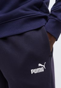Personne portant un pantalon de survêtement Puma bleu marine et un sweat-shirt assorti, avec une main glissée dans la poche du pantalon de survêtement.
