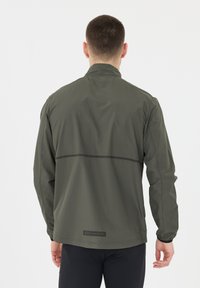 Man sett från bakom i en olivgrön lättviktsjacka med "ENDURANCE"-logotypen på nedre delen av ryggen och svarta träningsbyxor.